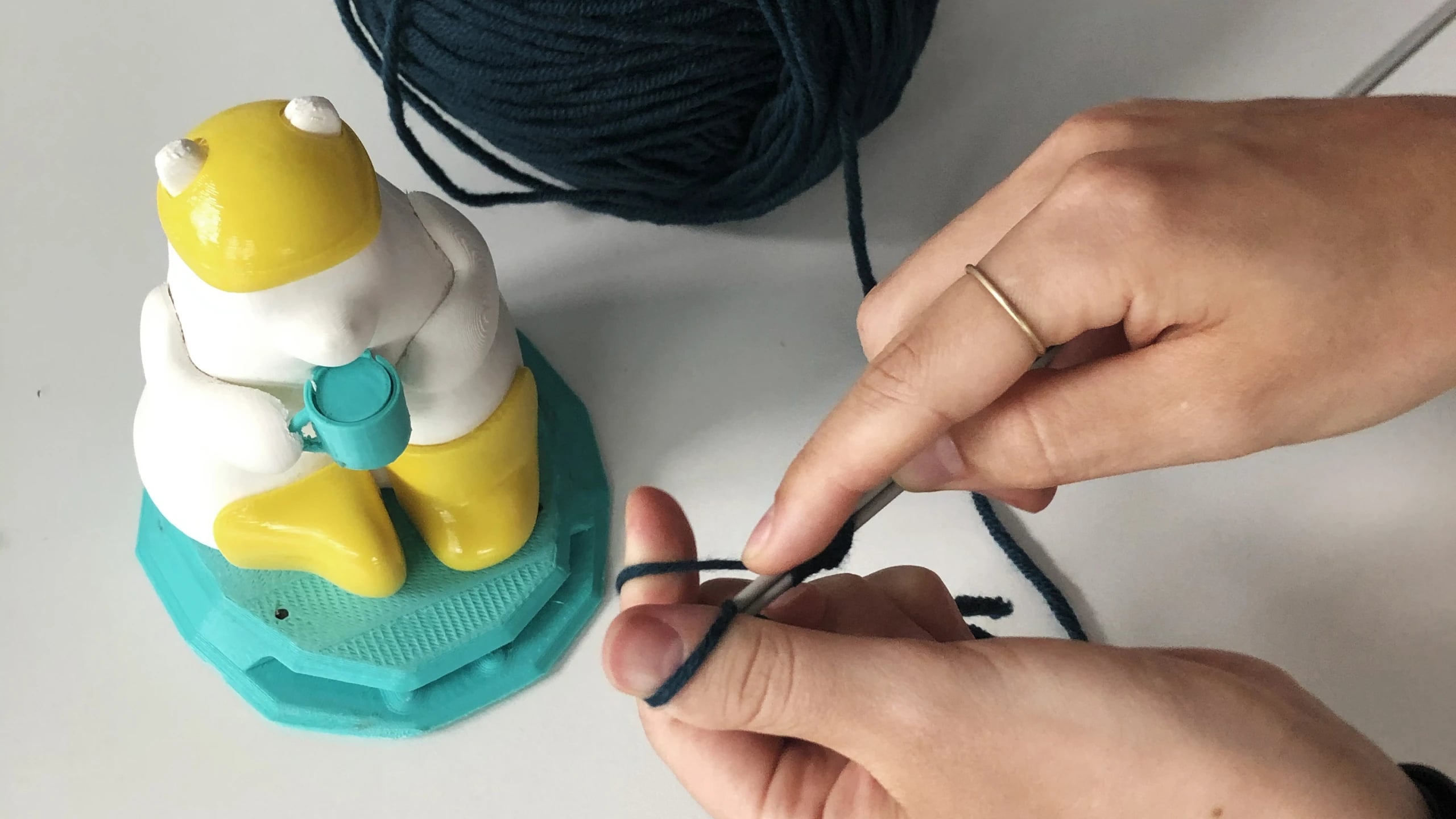 Hands knitting a navy scarf in fine yarn beside the painted Milo model on its turquoise base