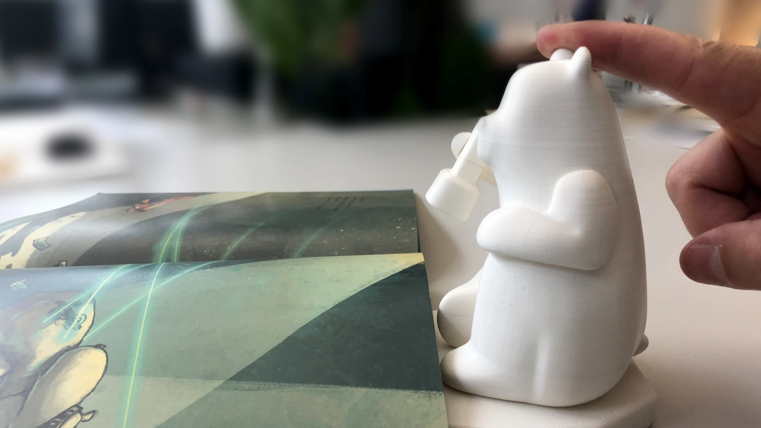 A finger pressing the back of a white 3D-printed Milo beside an open book, testing the speaker placement
