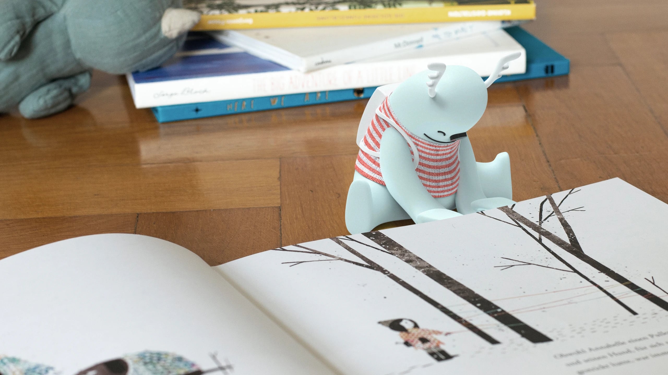 The Kit prototype, a pale blue antlered monster in a red-striped vest, perched on the edge of an open picture book