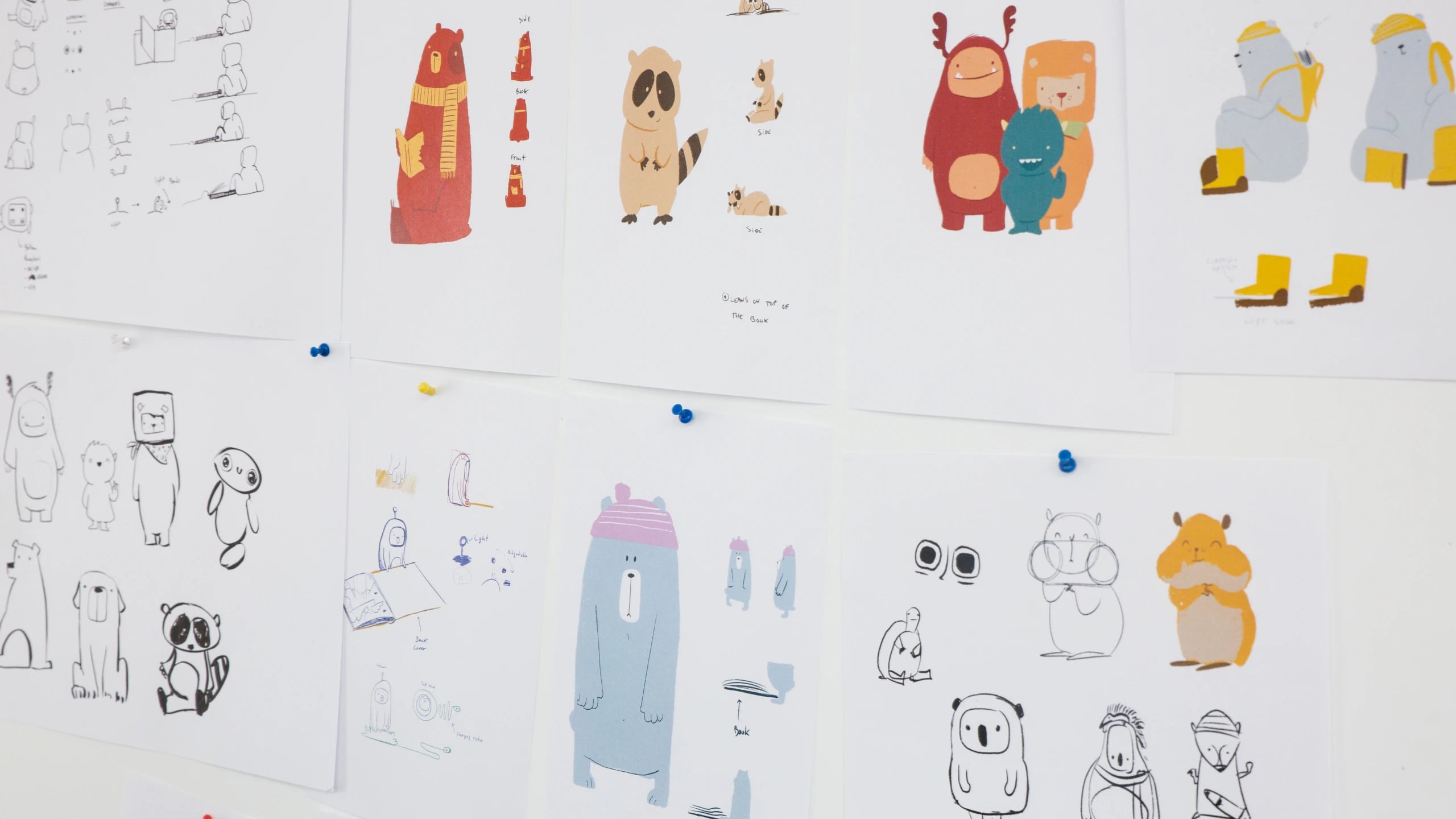 Character studies pinned to a studio wall, pencil roughs beside coloured drawings of bears, raccoons, monsters and hamsters
